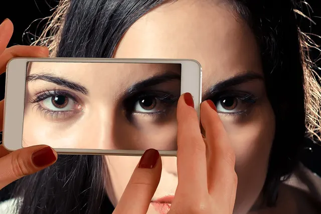 Hands hold a white smartphone in front of a dark-haired woman's face, the screen showing a close-up of her eyes