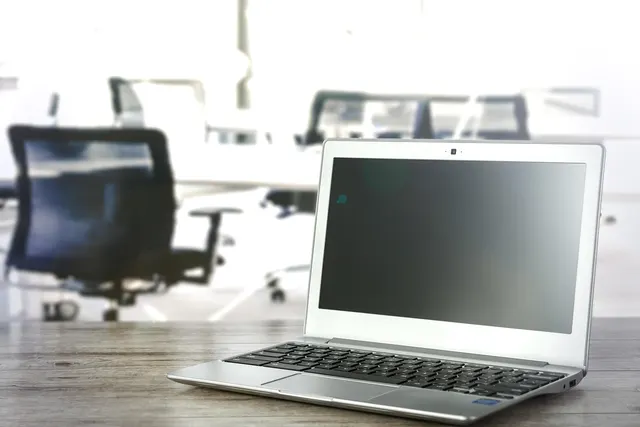 Silver laptop with a blank screen on a wooden desk in a bright office with blurred black chairs behind it