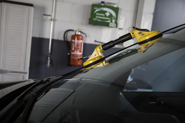 Yellow suction cup tool on a car windshield in an auto repair bay, fire extinguisher and first aid kit on wall