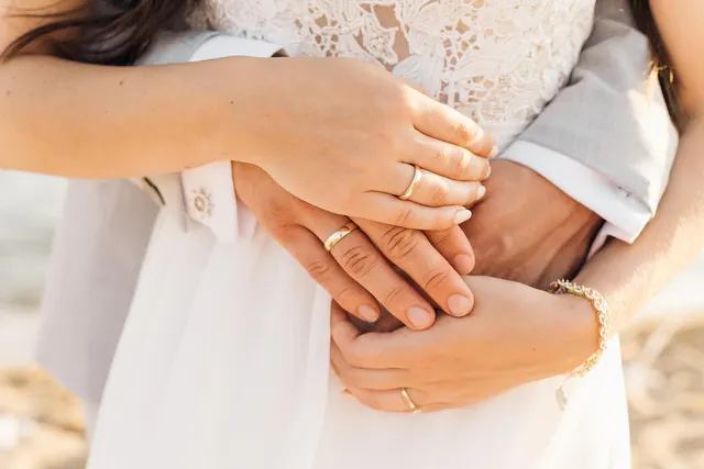 Close-up of a couple embracing, hands stacked to show gold wedding bands, bride in a white lace dress