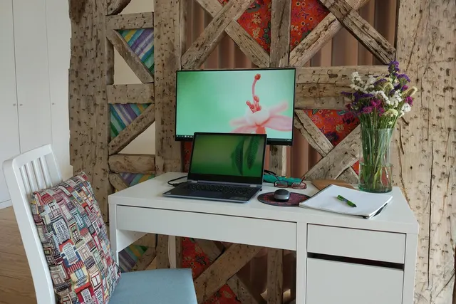 Home office desk with a laptop, monitor, notebook and vase of wildflowers against a rustic timber-framed wall
