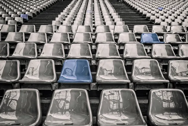 Rows of empty gray plastic stadium seats with a few blue seats scattered among them, rising up the stands