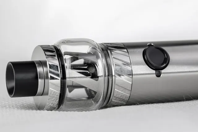 Close-up of a silver vape pen with a clear glass tank, black drip tip and fire button on a white fabric surface