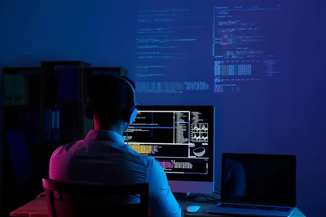 Developer wearing headphones works at a desktop monitor showing code in a dark office lit by blue and purple light