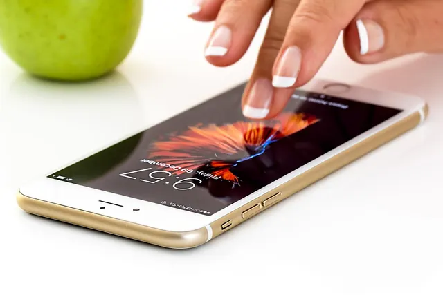 Manicured finger tapping a gold smartphone lock screen showing 9:57 on a white desk, green apple blurred behind