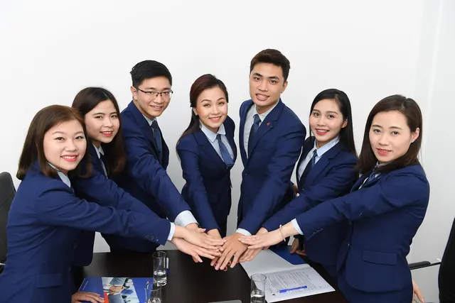 Seven smiling staff in matching navy suits and ties stack hands over a conference table with documents and water glasses