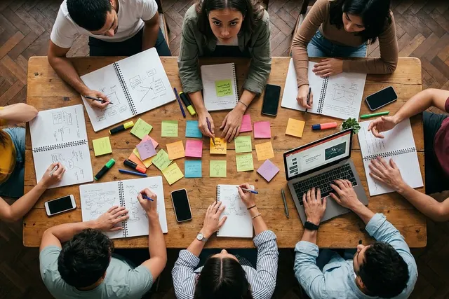 Overhead view of eight colleagues planning at a wooden table with notebooks, colorful sticky notes, phones and a laptop