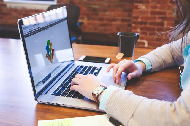 Woman typing on a laptop at a wooden desk with a smartphone, mug and notepad, brick wall in the background