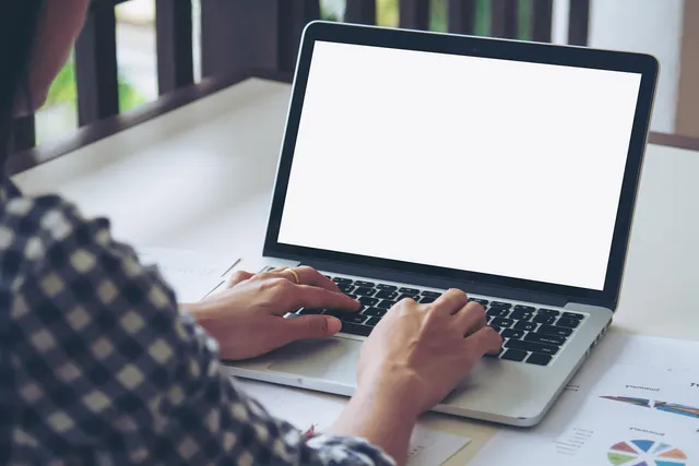 Person in a checkered shirt typing on a laptop with a blank white screen, with printed charts on the desk