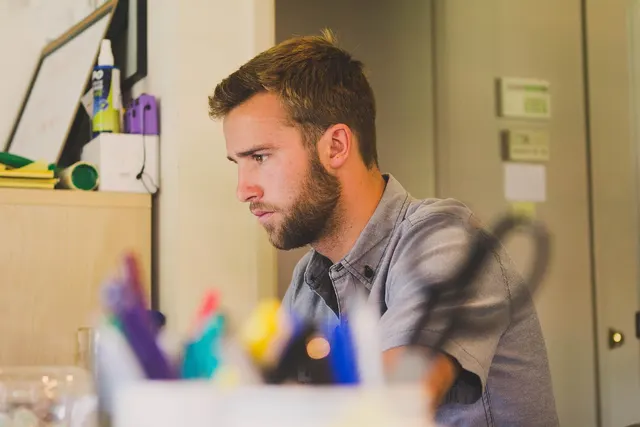 Bearded man in a gray shirt concentrating at his desk in a small office, with a cup of colorful markers in the foreground