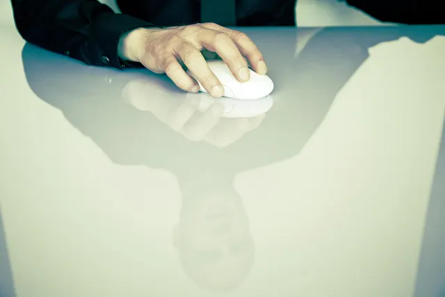 Hand in a dark suit sleeve resting on a white computer mouse, reflected with the man's face in a glossy white desk