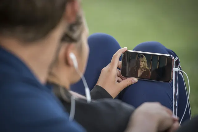 Person with white earbuds watching a streaming video on a smartphone held sideways, sitting outdoors on grass