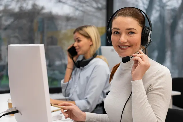 Smiling support agent in a headset at a computer while a colleague takes a phone call behind her in a bright office