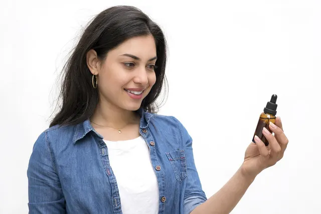 Smiling woman in a denim shirt holding up an amber glass dropper bottle of supplement against a white background