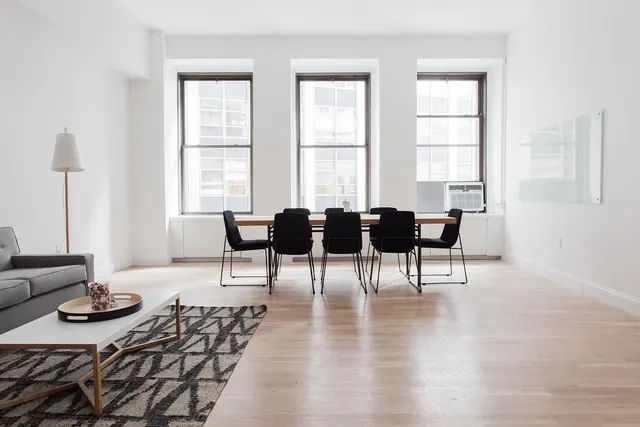 Bright furnished room with gray sofa, white coffee table on patterned rug, and wooden dining table with black chairs