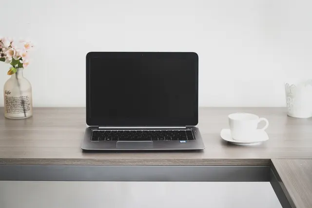 Silver laptop with a blank screen on a wooden desk beside a white coffee cup, with pink flowers in a vase