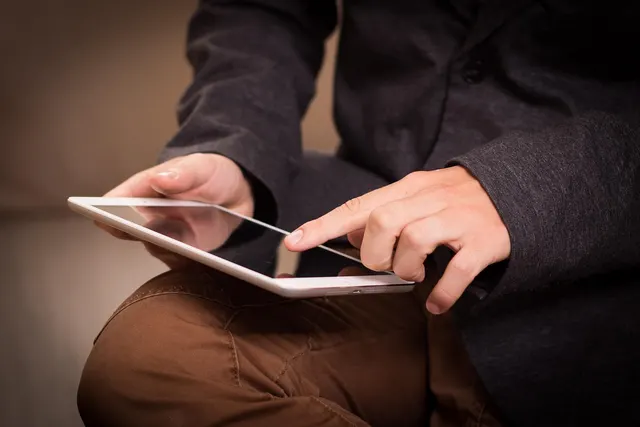 Close-up of a seated man in a dark gray blazer and brown trousers tapping the screen of a white tablet