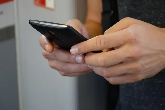 Close-up of a person's hands holding and tapping a black smartphone against a blurred light background