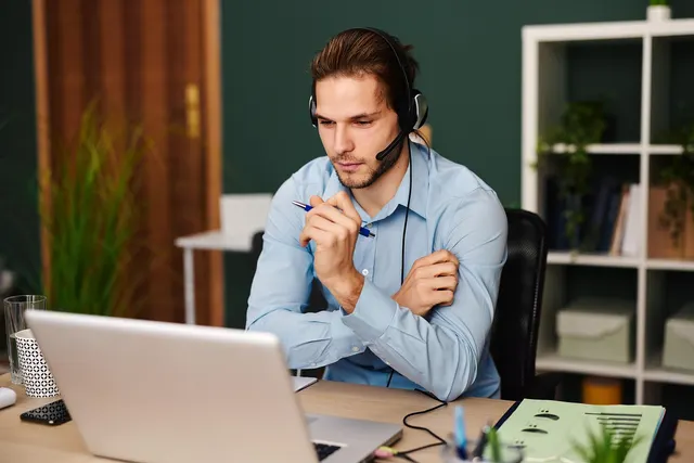 Man in a light blue shirt wearing a headset, holding a pen and looking at a laptop while taking a call at an office desk
