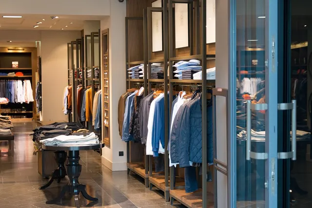 Clothing boutique interior with jackets and shirts on wooden racks, folded garments on a table, and glass entry doors