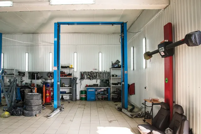 Empty auto repair garage with a blue two-post car lift, stacked tires, tool wall, red alignment rig and removed car seats