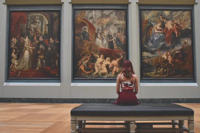 Woman with red hair sits on a bench in a museum gallery, viewing three large Baroque paintings on the wall