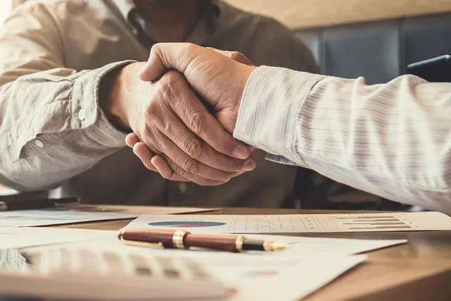 Two professionals shake hands over a wooden desk covered with financial charts, a calculator and a gold-trimmed pen