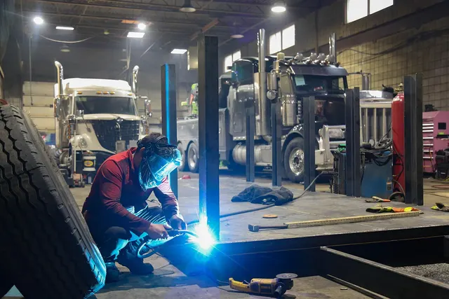 Mechanic in a welding mask welding steel posts on the floor of a heavy truck repair shop, with semi trucks behind