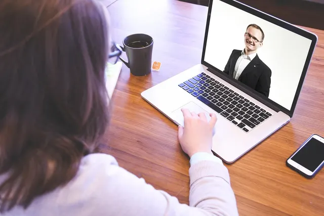 Woman at a wooden desk on a laptop video call with a smiling man in a suit, beside a coffee mug and smartphone