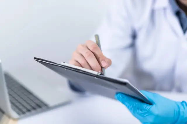 Gloved clinician writing on a clipboard beside a laptop