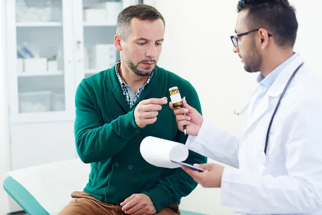 Doctor in white coat holding a clipboard hands a prescription pill bottle to a male patient in an exam room
