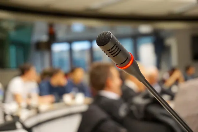 Close-up of a conference microphone with a red ring, with blurred attendees seated in a meeting room behind it