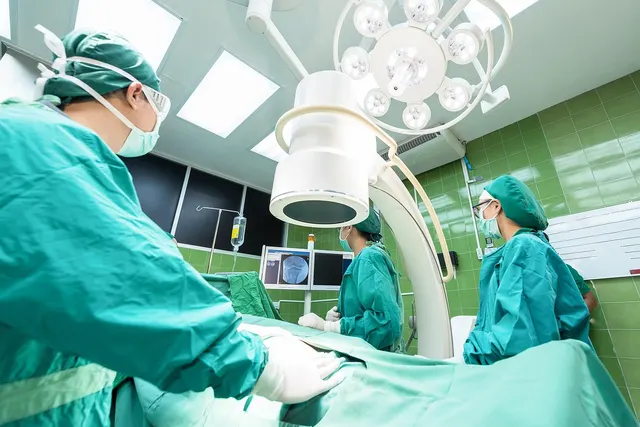 Surgical team in green scrubs, caps and masks operating under bright lights with a C-arm and imaging monitor
