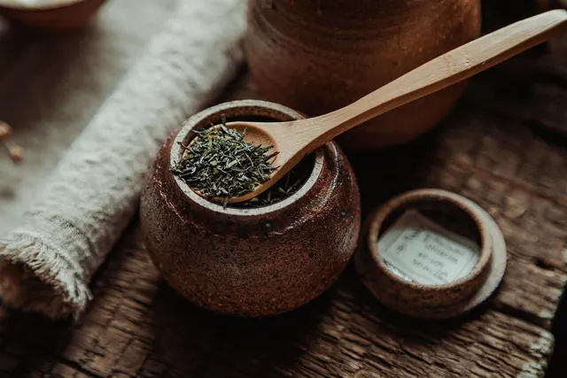 Dried crushed green leaves in a rustic ceramic jar with a wooden spoon, beside a small lid and linen cloth