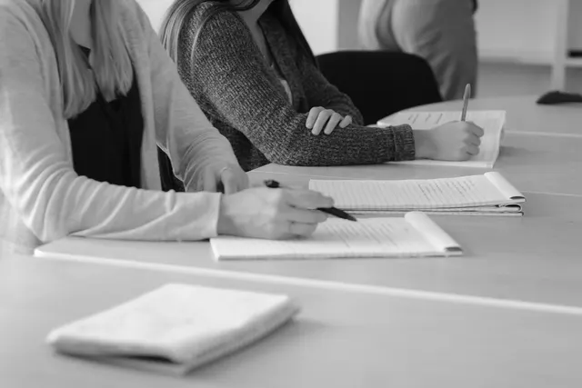 Black and white photo of two people writing notes in notepads at a shared table during a meeting