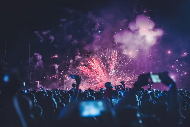 Crowd at a night concert holding up phones as red fireworks burst amid purple smoke above the stage
