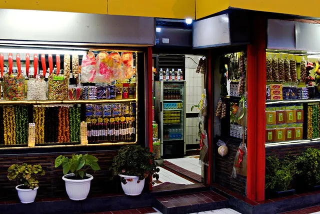 Brightly lit convenience store front with windows stocked with packaged snacks and candy, potted plants outside