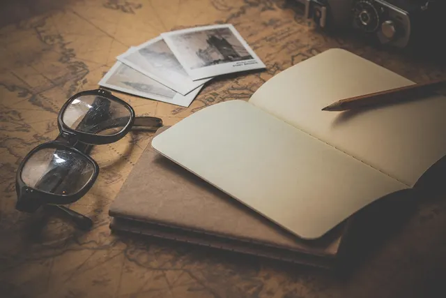 Open blank notebook with pencil, eyeglasses, vintage photos and a camera on an antique world map