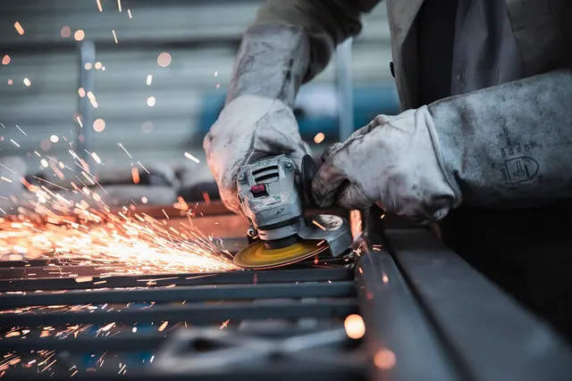 Gloved metalworker cutting a steel grate with an angle grinder, orange sparks flying in a workshop