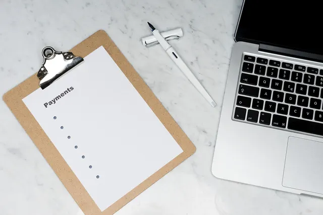 Clipboard with a blank Payments checklist beside a white pen and an open laptop on a white marble desk