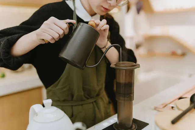 Barista in a green apron pouring water from a gooseneck kettle into a coffee press on a scale at a café counter