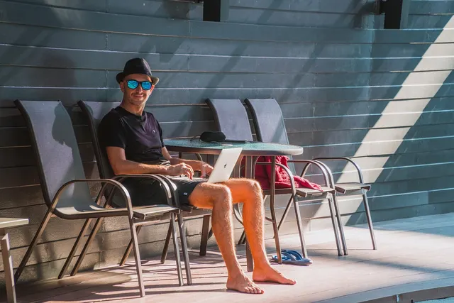 Barefoot man in a hat and sunglasses works on a laptop at a table on a sunny poolside deck at a vacation property