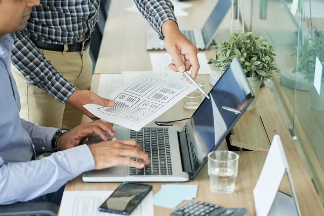 Two colleagues review a paper wireframe sketch beside a laptop, smartphone and calculator on an office desk