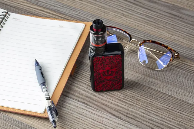 Black vape mod with red damask panel and tank beside eyeglasses, an open lined notebook and a pen on a wooden desk