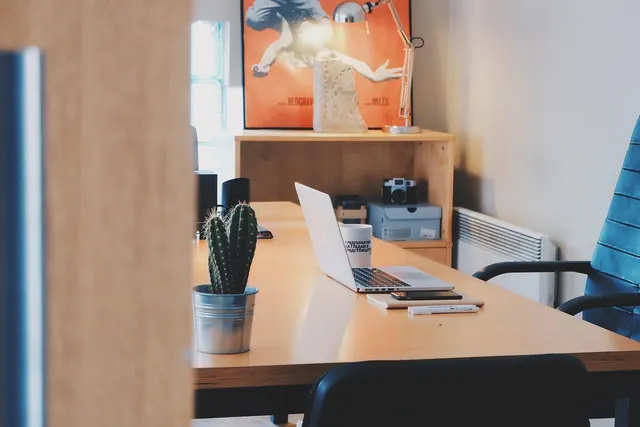 Sunlit office desk with an open laptop, smartphone, notebook and potted cactus beside a blue office chair