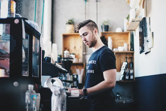 Barista at a coffee shop counter reading a printed receipt beside a POS terminal and receipt printer