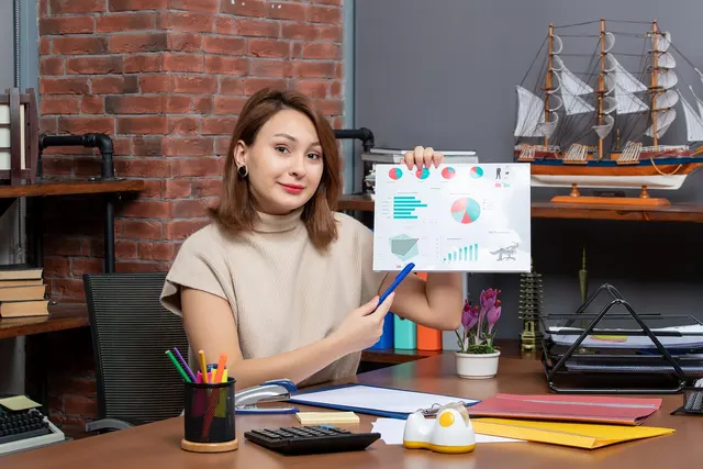 Woman at an office desk holds up a printed report of charts and points to it with a pen, beside a calculator and folders