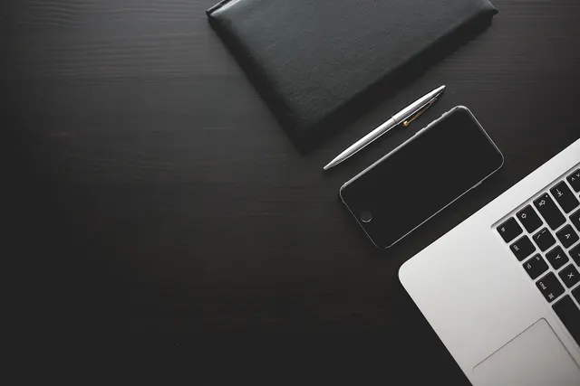 Overhead view of a dark wooden desk with a black leather notebook, silver pen, smartphone and the corner of a laptop