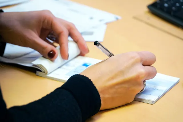 Close-up of a woman's hands writing in a checkbook with a ballpoint pen on a wooden office desk beside papers