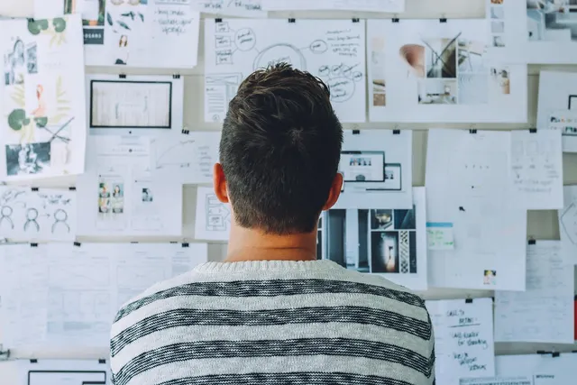 Man in a striped sweater, seen from behind, studying a wall of pinned website wireframes, sketches and design mockups